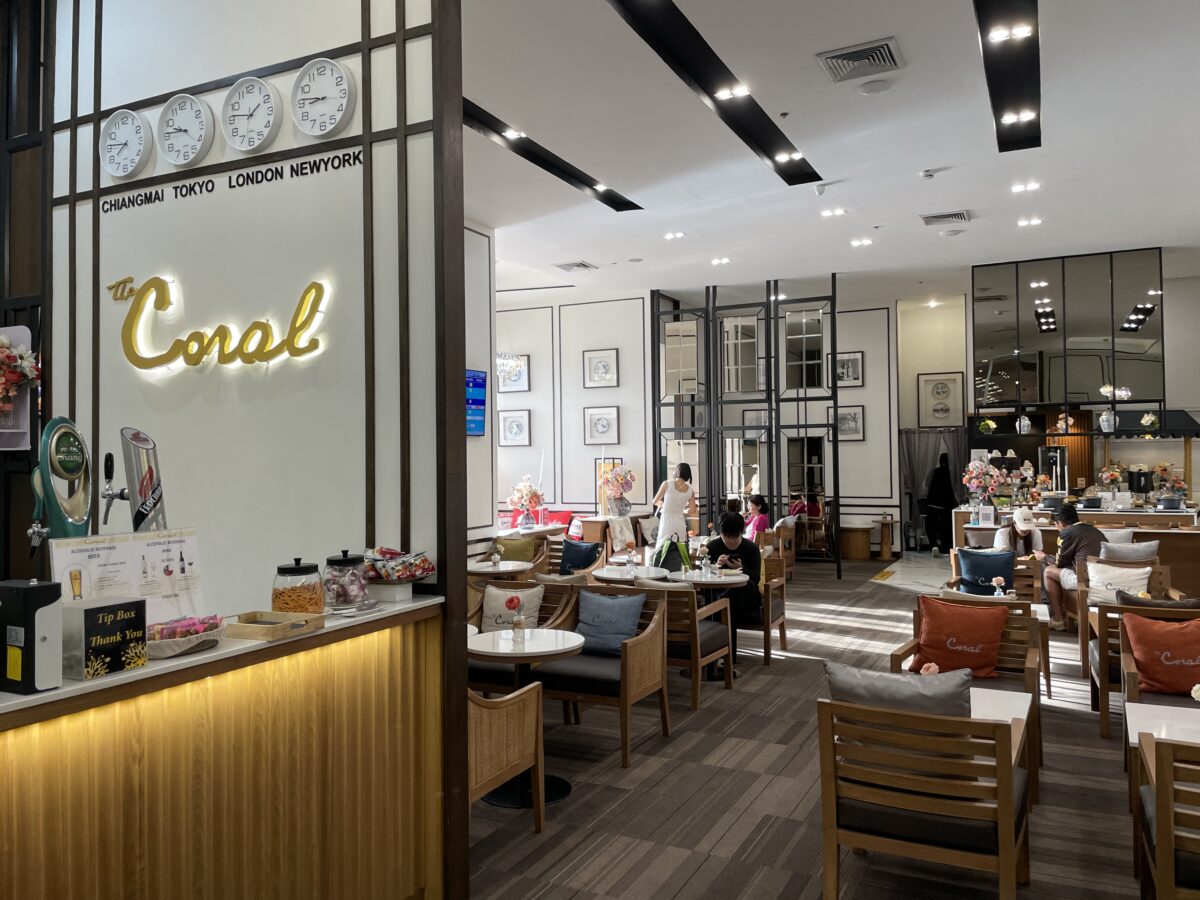 Coralの金色の看板と各都市の時計を掲げたラウンジの入口。奥に木の椅子とテーブルの席が広がる